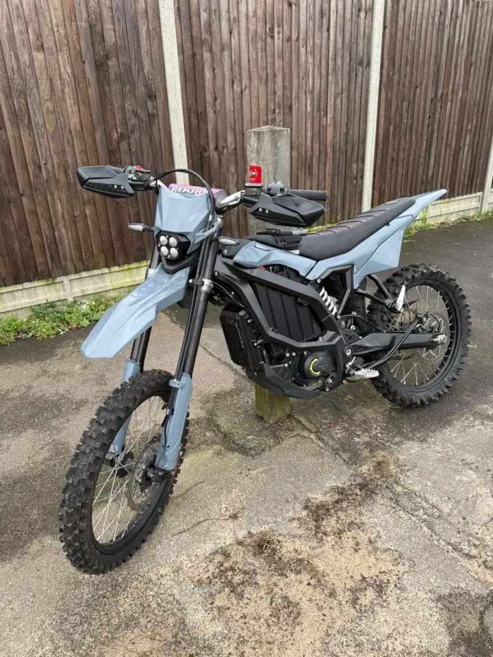 Grey electric dirt bike with a four-lens LED headlight, hand guards and a Renthal bar pad, on a paved path
