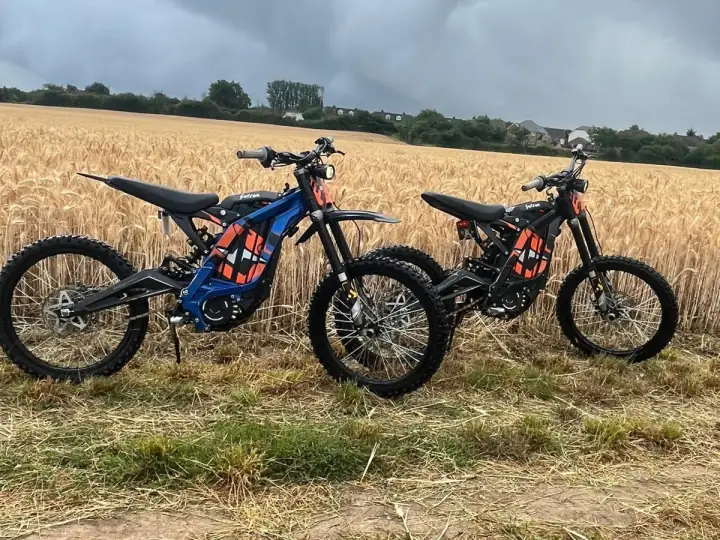 Blue and black Sur-Ron electric dirt bikes parked at the edge of a wheat field under a stormy sky